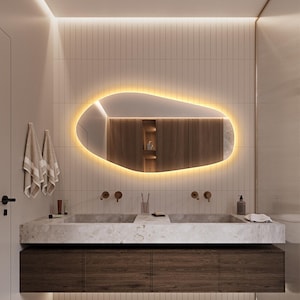 May include: Modern bathroom design featuring a large, uniquely shaped mirror with warm lighting. The mirror is above a double sink with a marble countertop and dark wood cabinetry. The walls are tiled, and the room has a minimalist aesthetic.
