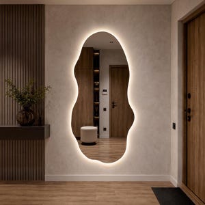 May include: A large, irregular, organic-shaped wall mirror featuring a smooth, curved edge and integrated backlighting that emits a soft, warm glow around its perimeter, creating a distinct silhouette against the surrounding wall surface.