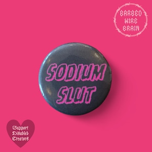 Può includere: Un bottone nero con il testo "SODIUM SLUT" in lettere rosa. Il bottone è su uno sfondo rosa.