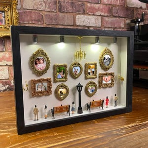 Może przedstawiać: Gablota z miniaturowymi oprawionymi zdjęciami, latarnią uliczną, ławkami i maleńkimi figurkami. Ramki są złote i w różnych kształtach. Tło to ceglana ściana.