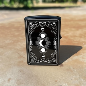 May include: Black lighter with a white celestial design featuring crescent moons, suns, and stars. The design is framed by ornate, decorative borders. The lighter is rectangular and has a shiny finish.