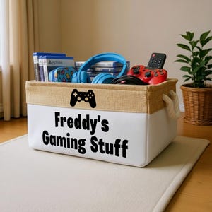 Może przedstawiać: Biały pojemnik do przechowywania z czarnym napisem "Freddy's Gaming Stuff". Wypełniony grami wideo, niebieskim zestawem słuchawkowym i czerwonym kontrolerem do gier. Pojemnik ma beżowe wykończenie z juty i uchwyty z liny.