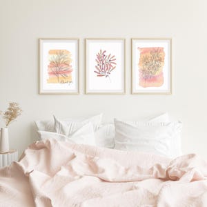 Puede incluir: Tres impresiones de acuarela enmarcadas con ilustraciones botánicas en una pared blanca sobre una colcha de cama rosa. Las impresiones presentan ramas delicadas con hojas y bayas en tonos de rosa y naranja.