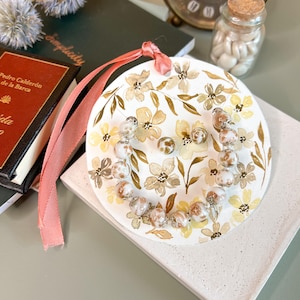 May include: A white round ornament with a floral pattern and a beaded necklace in shades of brown and white. The ornament is tied with a pink ribbon.