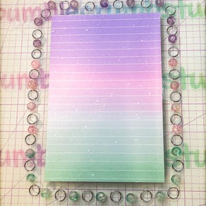 May include: A pastel pink, purple, and green lined notepad with a glittery finish. The notepad is surrounded by a border of silver rings with clear, glittery beads.