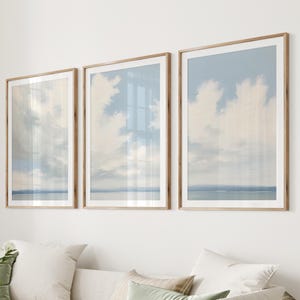 May include: Three framed art prints showing a tranquil sky and sea scene. The prints have a light blue and white colour scheme, with a wooden frame. The artwork is displayed above a white sofa with decorative cushions.