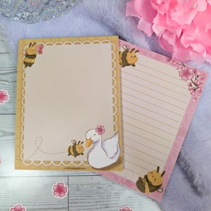May include: Two notepads with floral and bee illustrations. One notepad has a beige background with a scalloped border and a cartoon bee and duck. The other notepad has a pink border with floral accents and a cartoon bee. Pink flower stickers are scattered around.