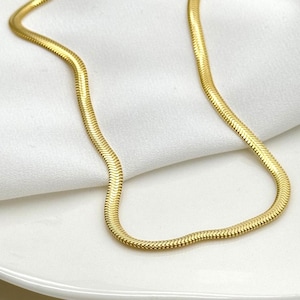Op de afbeelding: Een goudkleurige visgraat ketting. De ketting wordt gepresenteerd op een wit oppervlak. De ketting heeft een plat, flexibel ontwerp, met een subtiele glans. De ketting is een klassiek sieraad.