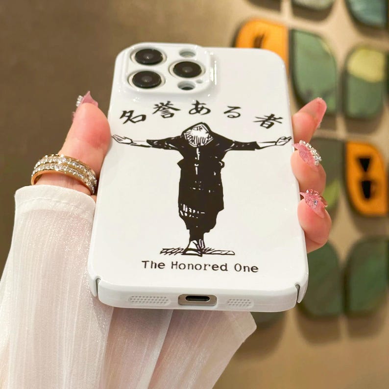 May include: White phone case featuring a black illustration of a figure with outstretched arms. The case has Japanese text and the English text "The Honored One". The case has a camera cutout and button holes.