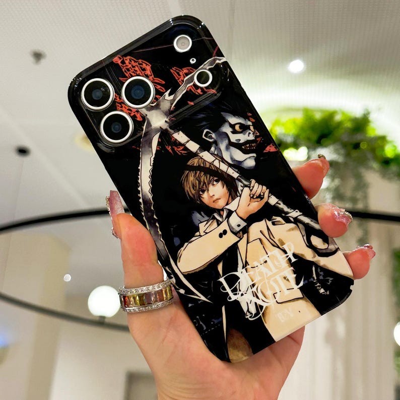 May include: A black smartphone case featuring a graphic illustration of the characters Light Yagami and Ryuk from Death Note. The design includes a large scythe, the Death Note logo in white text, and a glossy finish with cutouts for the camera lenses.