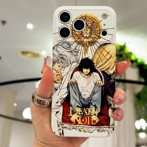 May include: A white smartphone case featuring a graphic illustration of characters from Death Note, including L sitting on a red chair. The design includes a gold circular emblem at the top and the text Death Note in gold lettering at the bottom.