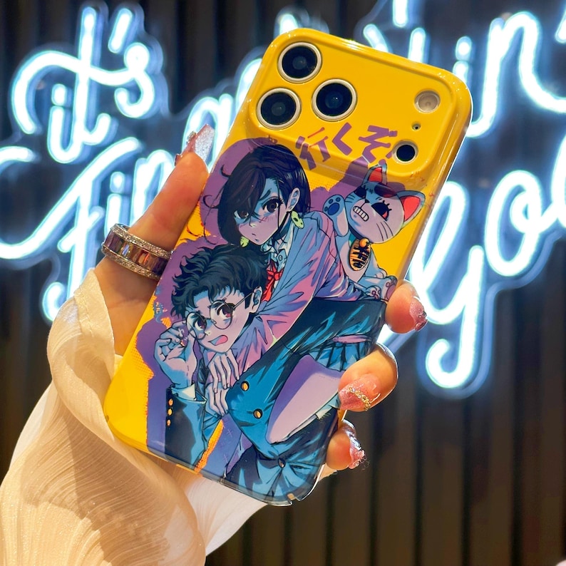 May include: Yellow phone case featuring an anime-style illustration of two characters and a cat. The artwork is in shades of blue, purple, and pink. The case has a glossy finish and a camera cutout.
