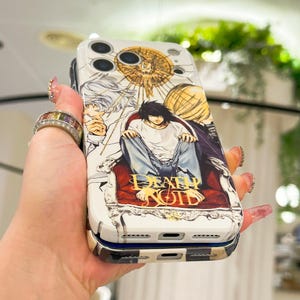 May include: A white smartphone case featuring a graphic illustration of characters from Death Note, including L sitting on a red chair. The design includes gold circular accents and the Death Note logo in gold lettering at the bottom of the case.