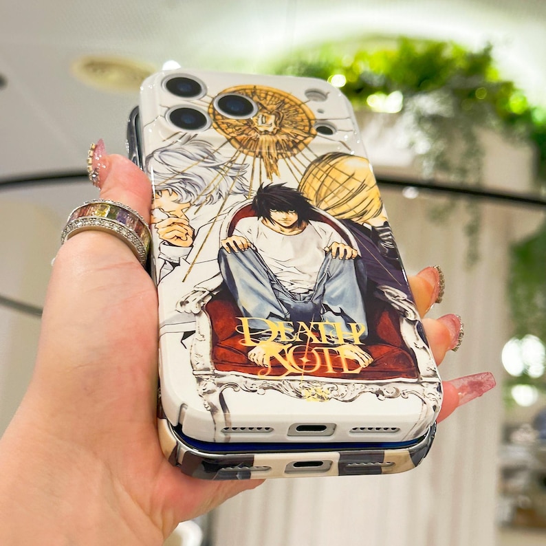 May include: A white smartphone case featuring a graphic illustration of characters from Death Note, including L sitting on a red chair. The design includes gold circular accents and the Death Note logo in gold lettering at the bottom of the case.