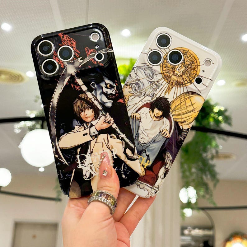 May include: Two smartphone cases featuring anime-style illustrations. The black case displays a character holding a scythe with the text Death Note. The white case features two characters with a golden circular emblem above them. Both cases have camera cutouts.