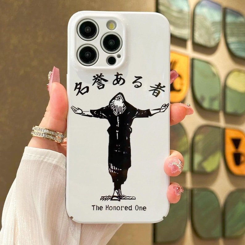 May include: White phone case with a black graphic of a figure with outstretched arms, and Japanese text above. The text "The Honored One" is printed below the figure. The case has a camera cutout with three lenses.