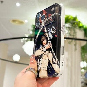 May include: A black and white smartphone case featuring a graphic illustration of the characters Light Yagami and Ryuk from Death Note. The design includes the Death Note logo in white text, with a glossy finish and cutouts for the camera lenses.