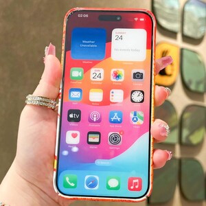 K&ouml;nnte beinhalten: Ein Smartphone mit einer rot-orangenen H&uuml;lle, das den Startbildschirm mit App-Symbolen anzeigt. Der Bildschirm zeigt das Datum Sonntag, den 24. und die Uhrzeit 02:06 Uhr an. Das Telefon wird in einer Hand mit einem Ring gehalten.