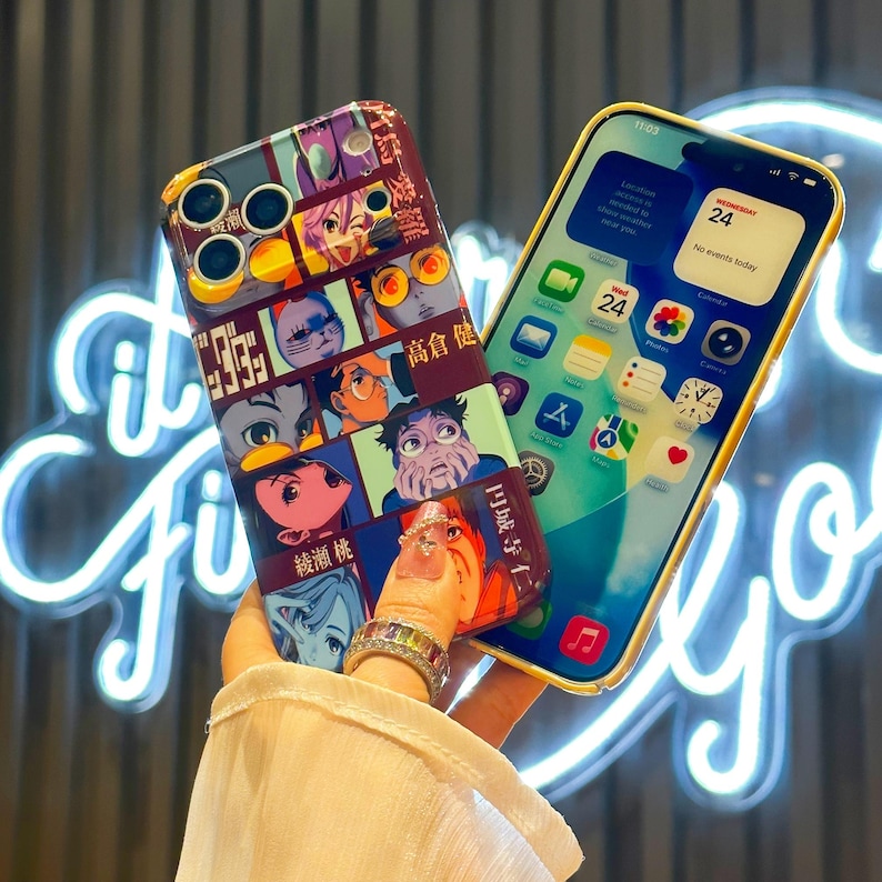 May include: A smartphone with a yellow case displays a home screen with app icons. Beside it, a phone case with a colorful anime-style design featuring various character faces and Japanese text. The case is held in a hand.