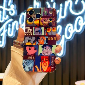 May include: A colorful phone case featuring a collage of anime-style illustrations. The case has a dark red background with multiple panels, each displaying a different character or scene in vibrant colors. The case has cutouts for the phone's camera and buttons.