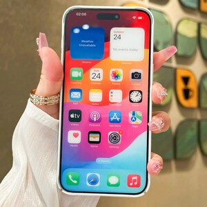 May include: A smartphone with a white case displays a colorful home screen with app icons. The screen shows the date as Sunday, the 24th, and the time as 02:06. The phone is held in a hand with a gold ring.