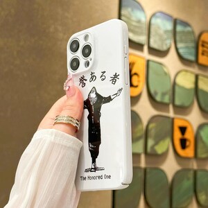 May include: White phone case featuring a black and white illustration of a character with outstretched arms. The case has Japanese text and the English text "The Honored One". The case has a glossy finish and a camera bump.
