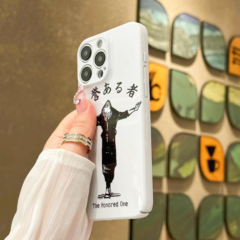 May include: White phone case featuring a black and white illustration of a character with outstretched arms. The case has Japanese text and the English text "The Honored One". The case has a glossy finish and a camera bump.