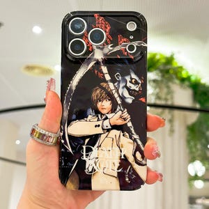 May include: A black smartphone case featuring a graphic illustration of a character with light brown hair holding a large scythe, accompanied by a dark, grinning figure. The text Death Note is printed in white at the bottom of the case.
