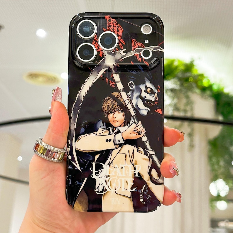 May include: A black smartphone case featuring a graphic illustration of a character with light brown hair holding a large scythe, accompanied by a dark, grinning figure. The text Death Note is printed in white at the bottom of the case.