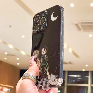 K&ouml;nnte beinhalten: Schwarze Handyh&uuml;lle mit einer Anime-Illustration von zwei Charakteren und einem Halbmond. Die H&uuml;lle hat eine gl&auml;nzende Oberfl&auml;che und ein sch&uuml;tzendes Design mit einem Kameraausschnitt.