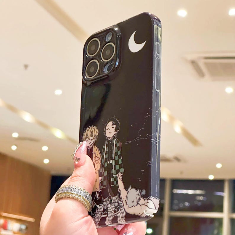 K&ouml;nnte beinhalten: Schwarze Handyh&uuml;lle mit einer Anime-Illustration von zwei Charakteren und einem Halbmond. Die H&uuml;lle hat eine gl&auml;nzende Oberfl&auml;che und ein sch&uuml;tzendes Design mit einem Kameraausschnitt.