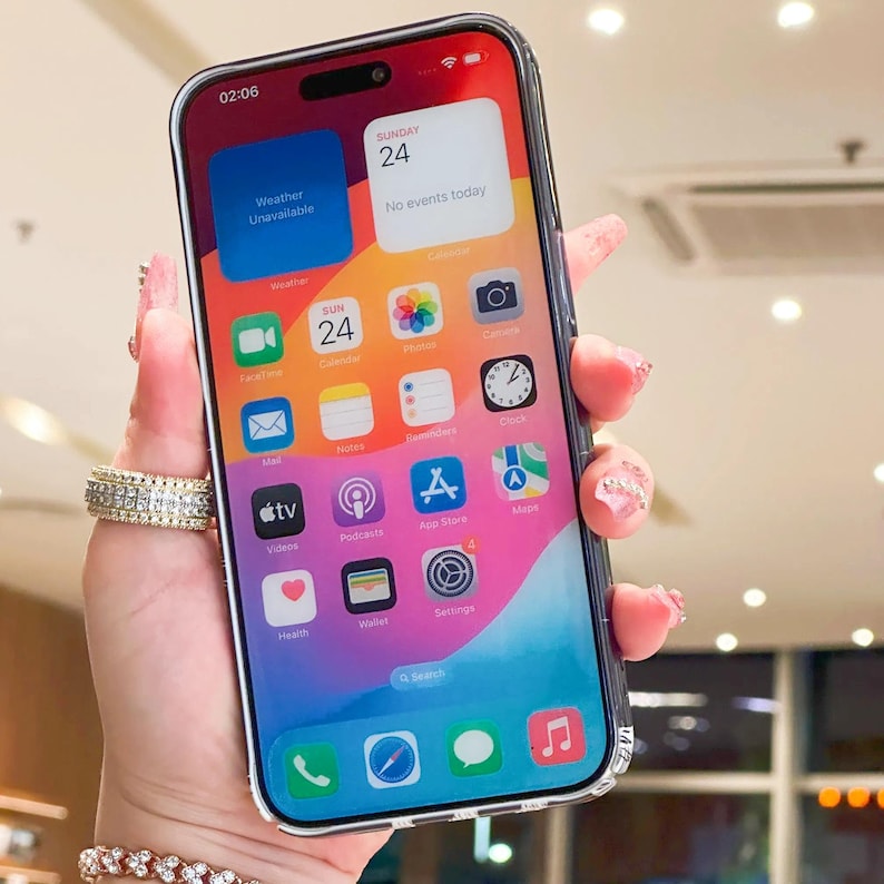 K&ouml;nnte beinhalten: Eine transparente Handyh&uuml;lle umschlie&szlig;t ein Smartphone, das einen farbenfrohen Startbildschirm mit App-Symbolen anzeigt. Der Bildschirm zeigt das Datum Sonntag, den 24. und die Uhrzeit 02:06 Uhr an. Das Telefon wird in einer Hand mit Ring und Armband gehalten.