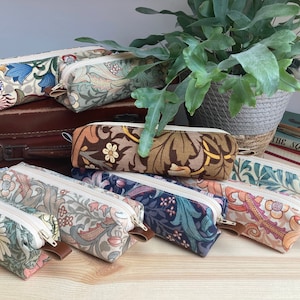 May include: A collection of eight colorful pencil cases with floral patterns. The cases are lined up on a wooden surface with a vintage suitcase and a potted plant in the background.