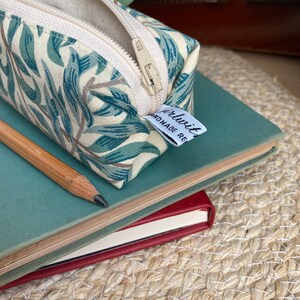 May include: A white and green floral patterned pencil case with a zipper closure. The case is made of fabric and has a small white tag attached to it that says "girlwith" and "HANDMADE RE".
