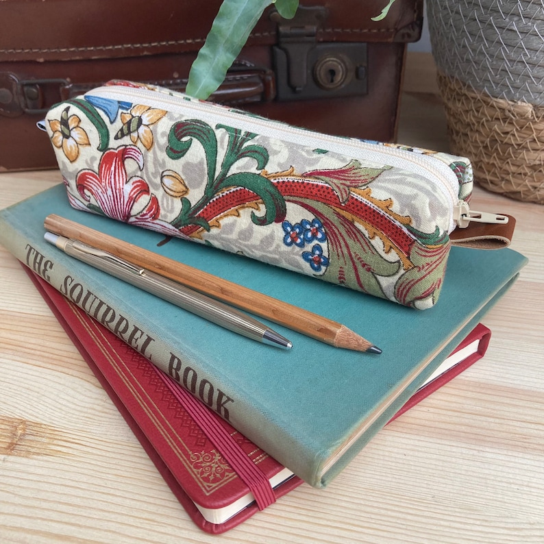 May include: A beige pencil case with a floral pattern in shades of red, green, and blue. The case is lying on top of two books, one blue and one red. The blue book has the title "The Squirrel Book" printed on the cover.
