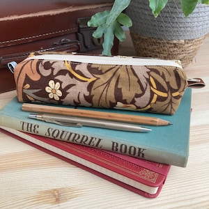 May include: A brown and beige pencil case with a floral pattern. The case is lying on top of a stack of books. The top book is blue and has the text "The Squirrel Book" printed on the cover. A silver and wooden pencil is lying on top of the books.