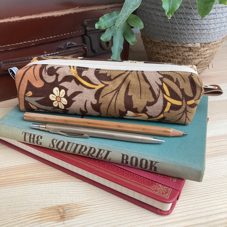 May include: A brown and beige pencil case with a floral pattern. The case is lying on top of a stack of books. The top book is blue and has the text "The Squirrel Book" printed on the cover. A silver and wooden pencil is lying on top of the books.