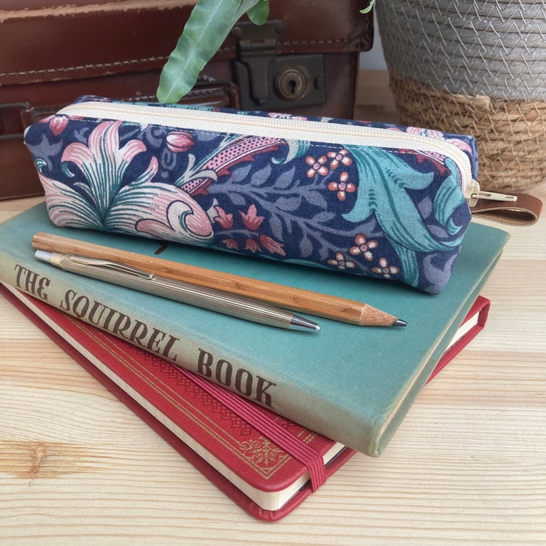 May include: A blue and pink floral pencil case with a zipper closure. The case is sitting on top of two books, one red and one green. The green book has the text "The Squirrel Book" on the cover.