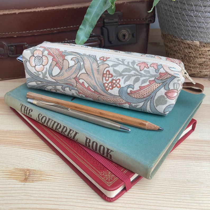 May include: A beige pencil case with a floral pattern in shades of pink, green, and blue. The case is lying on top of two books, one blue and one red. The blue book has the text "The Squirrel Book" printed on the cover.