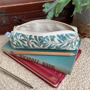 May include: A green and white pencil case with a floral pattern. The case is open and has a beige lining. The case is sitting on top of two books. The top book is blue and has the text "The Squirrel Book" printed on the cover. The bottom book is red and has gold lettering on the cover.