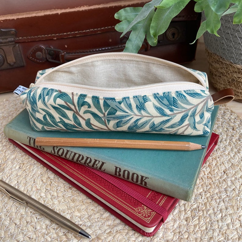 May include: A green and white pencil case with a floral pattern. The case is open and has a beige lining. The case is sitting on top of two books. The top book is blue and has the text "The Squirrel Book" printed on the cover. The bottom book is red and has gold lettering on the cover.