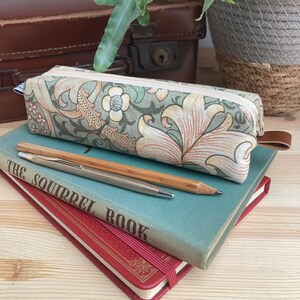 May include: A green and cream floral pencil case with a zipper closure. The case is sitting on top of two books, one blue and one red. The blue book has the text "The Squirrel Book" on the cover.