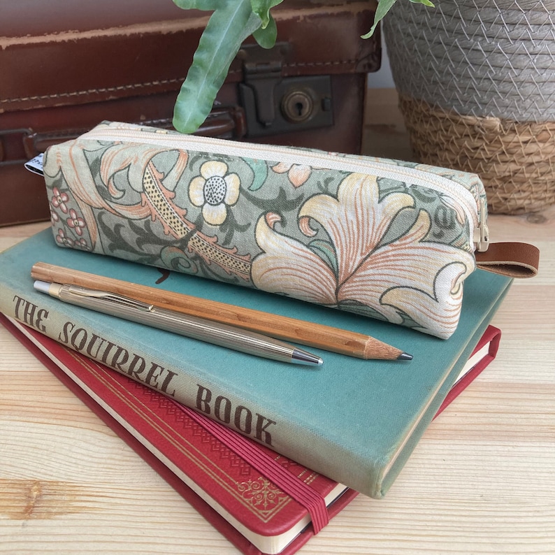 May include: A green and cream floral pencil case with a zipper closure. The case is sitting on top of two books, one blue and one red. The blue book has the text "The Squirrel Book" on the cover.
