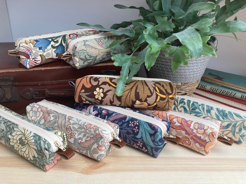 May include: A collection of eight colorful pencil cases with floral patterns. The cases are lined up on a wooden surface with a vintage suitcase and a potted plant in the background.