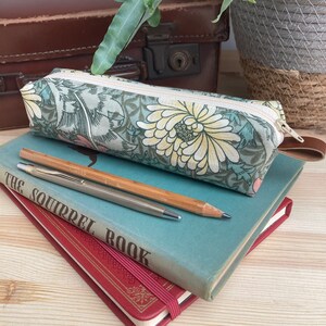 May include: A green and white floral pencil case with a zipper closure. The pencil case is sitting on top of two books, one red and one blue. A silver pen and a wooden pencil are on top of the books.