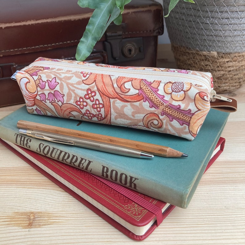 May include: A beige pencil case with a floral pattern in shades of orange, pink, and brown. The case is lying on top of a stack of books, one with the text "The Squirrel Book".