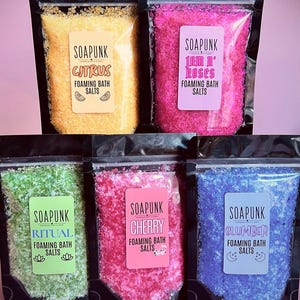 May include: Five black resealable pouches containing colourful foaming bath salts. The labels read Soapunk Citrus, Jam n' Roses, Ritual, Cherry, and Slumber. The salts are yellow, hot pink, light green, bright pink, and purple, respectively.
