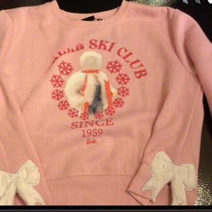 May include: Pink sweatshirt with a white bow on each sleeve. The front of the sweatshirt has a graphic of a snowman in a ski outfit with the text "Snow Ski Club Since 1959 Barbie".