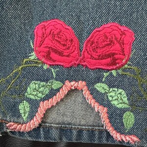 May include: A pair of red embroidered roses on a blue denim background. The roses are surrounded by green embroidered leaves and vines.