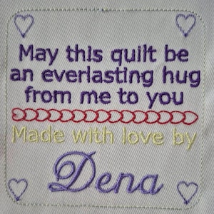May include: Purple embroidered text on a white fabric label reads "May this quilt be an everlasting hug from me to you Made with love by Dena". The text is surrounded by small hearts.